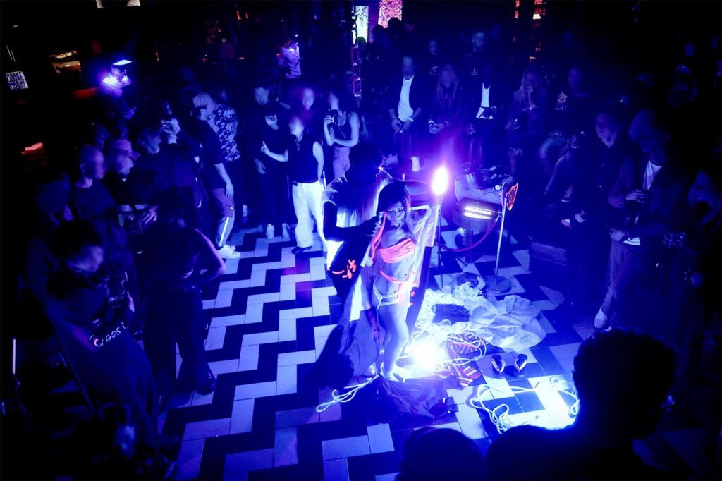 Shibari performance in a Tokyo nightclub by Louis Kordexe, showing a model illuminated by blue stage lighting while the audience gathers around the floor to watch.