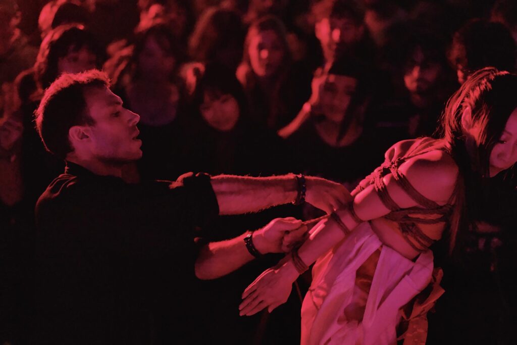Live shibari rope performance by Louis Kordexe in Tokyo, tying his model Kiyome on stage under red lighting with the audience watching.