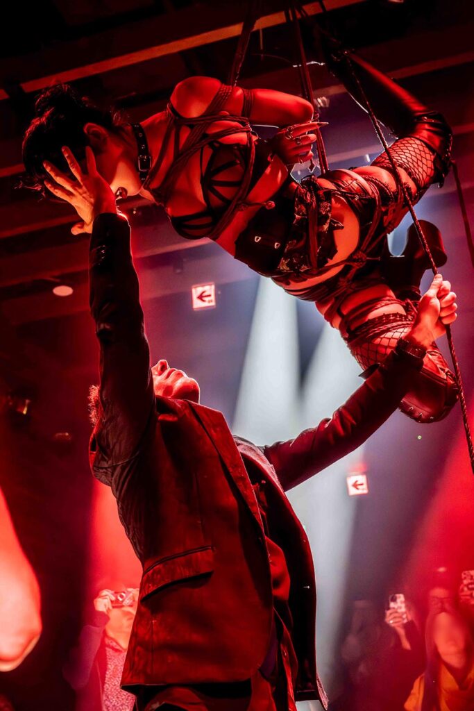 Shibari rope suspension performance by Louis Kordexe in Tokyo, featuring a model lifted on stage with red lighting while he supports her during the show.