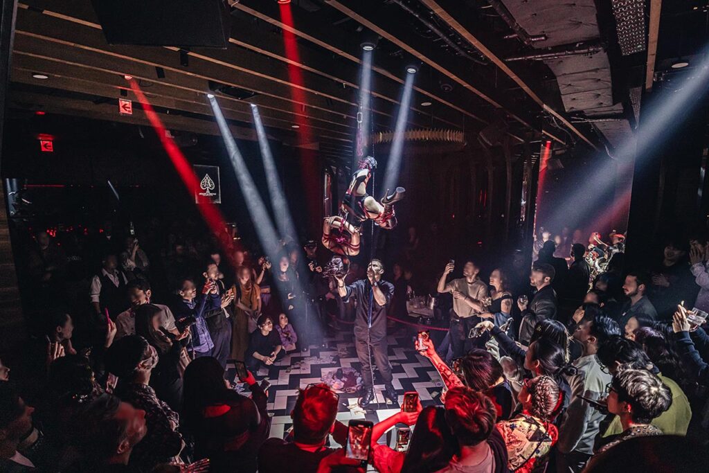 Shibari rope suspension performance by Louis Kordexe in a Tokyo nightclub, with a large audience surrounding the stage under dramatic spotlights.