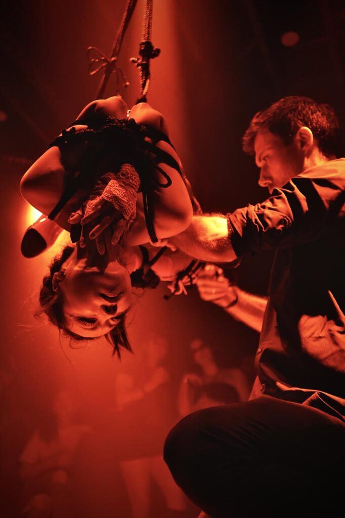 Live shibari rope performance in red lighting, featuring a dramatic stage with the performer adjusting the setup.