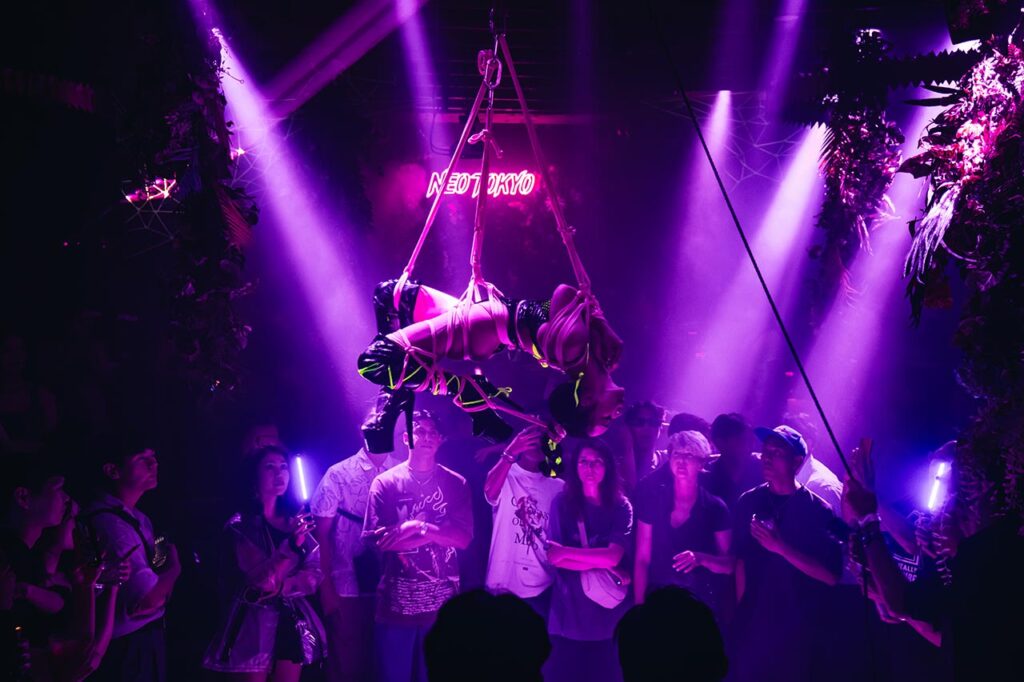 Neon UV shibari rope show by Louis Kordexe and Kiyome at a Tokyo nightlife event with a large crowd watching.